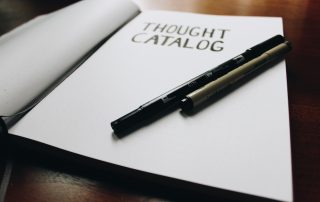 A notebook open on a table with the word 'though catalog' on. Two pens sit on top of the page.