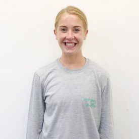 Jess facing the camera and smiling on a white background. She is wearing a grey top with the words Curious Motion on it in green.
