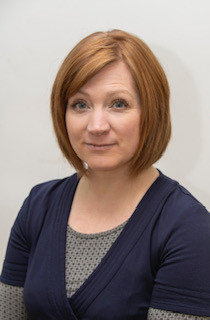 A photo of Kate Auker. She is wearing a dark v-neck t-shirt over a light grey long sleeved top, and she is smiling at the camera. Kate has white skin and red hair, which she wears in a bob.