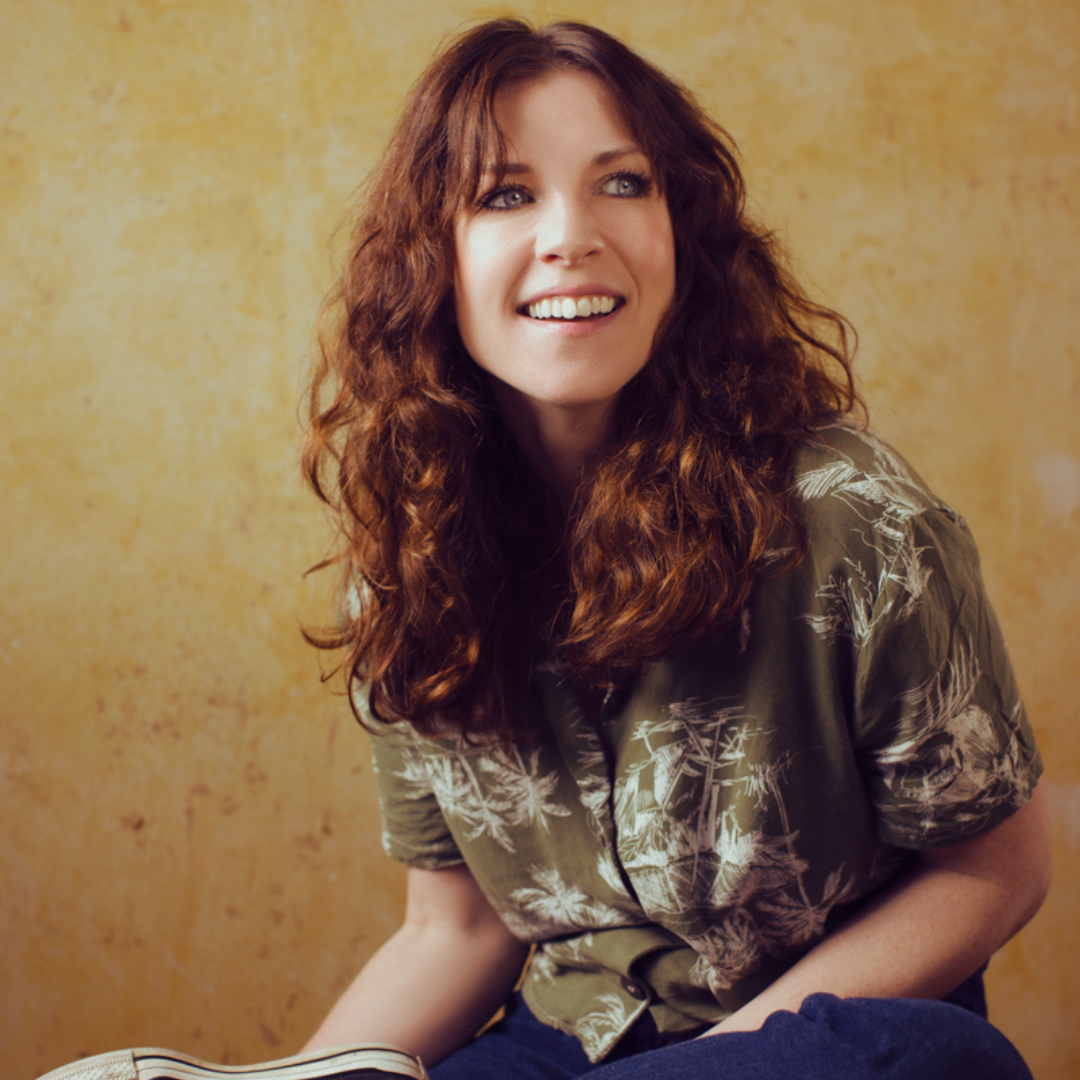 A photo of Rosie Doonan - a white female, with long curly auburn hair. She is smiling and looking up to the right corner.